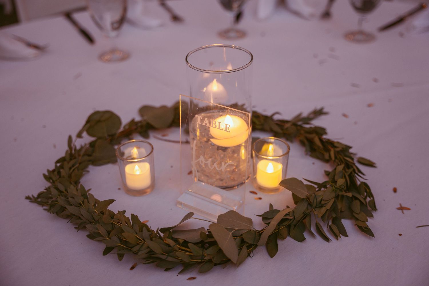 Glowing votive candles nestled in greenery wreaths create a romantic table setting with soft ambient lighting.
