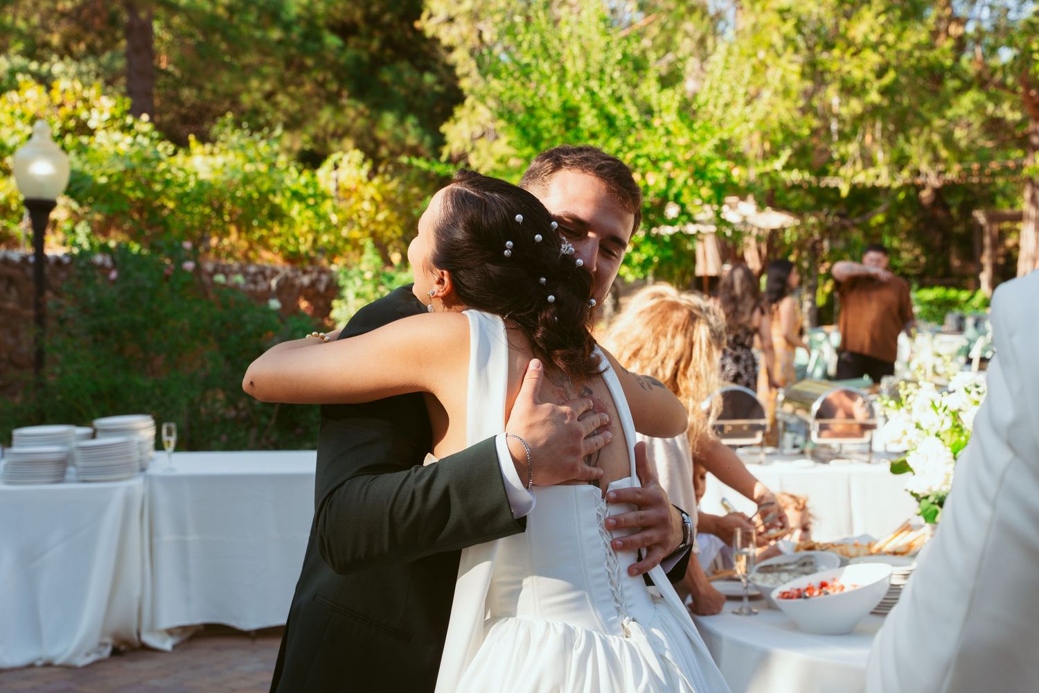 A couple embraces during an outdoor wedding reception with tables and guests in the background.