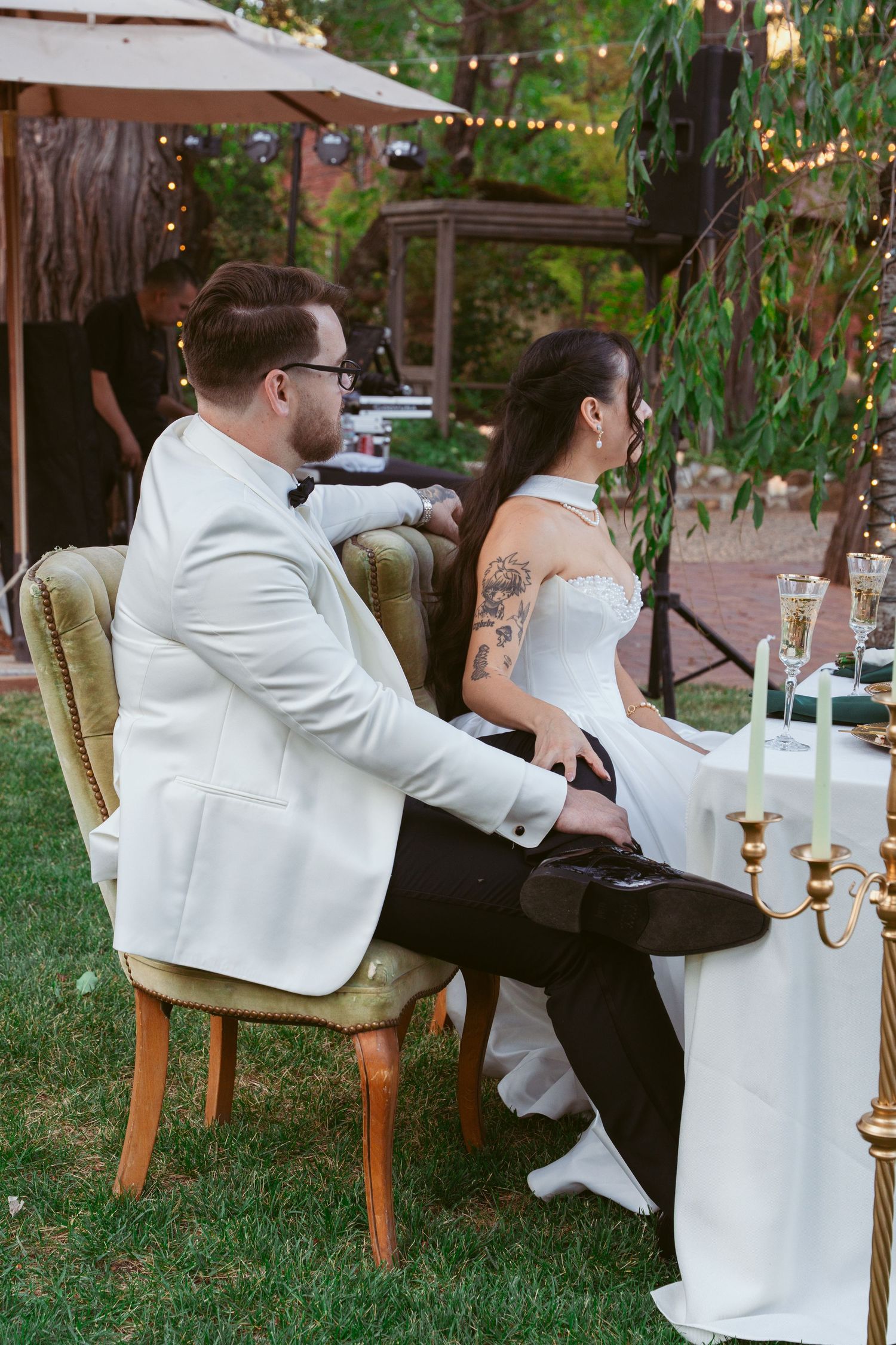 Couple sits together in white attire under string lights at an outdoor garden reception.