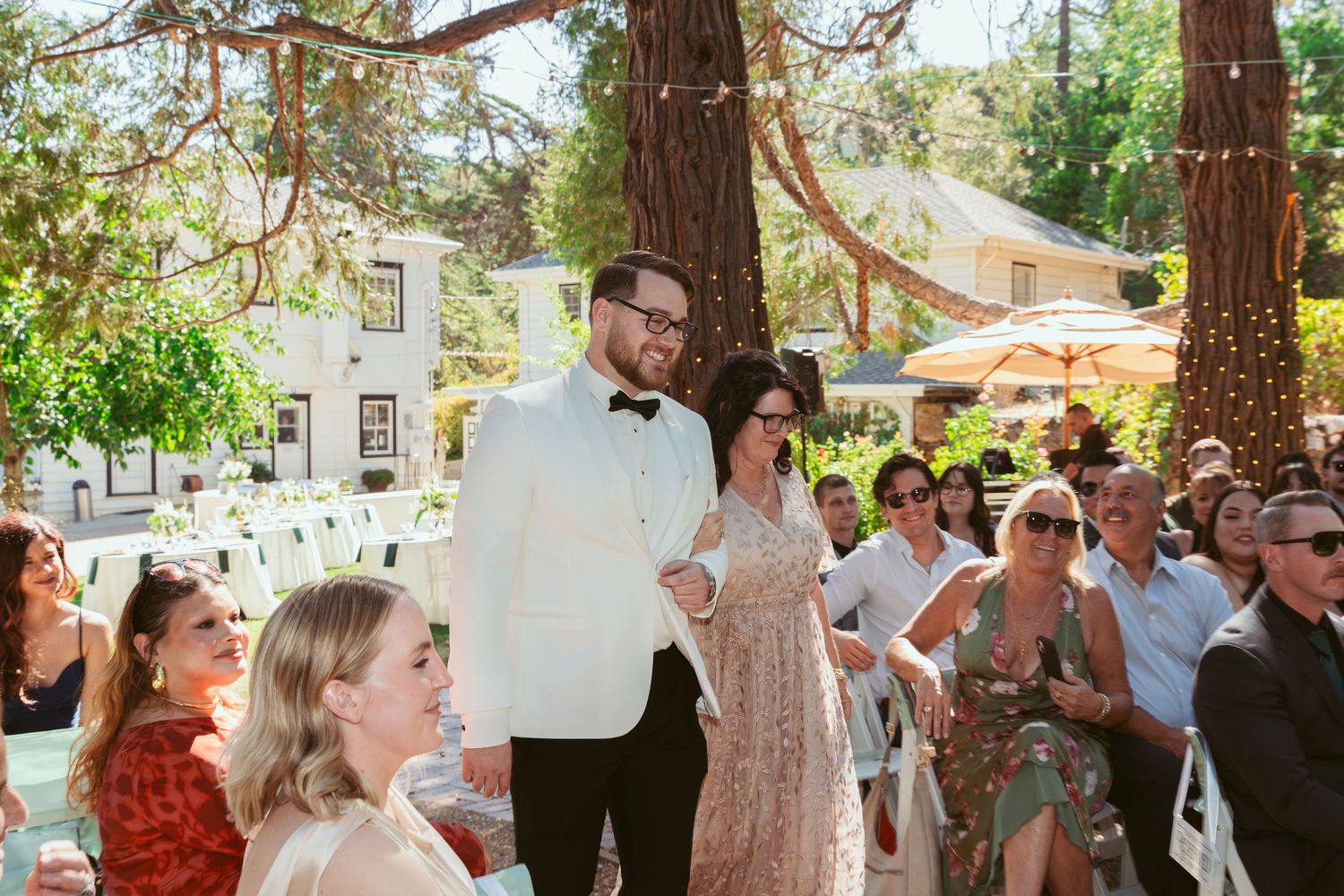 Outdoor wedding celebration in a garden setting with bunting decorations strung between tall pine trees.