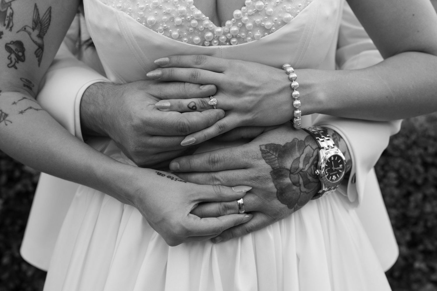 Close-up details of wedding rings and hands with delicate pearl accents on white dress.