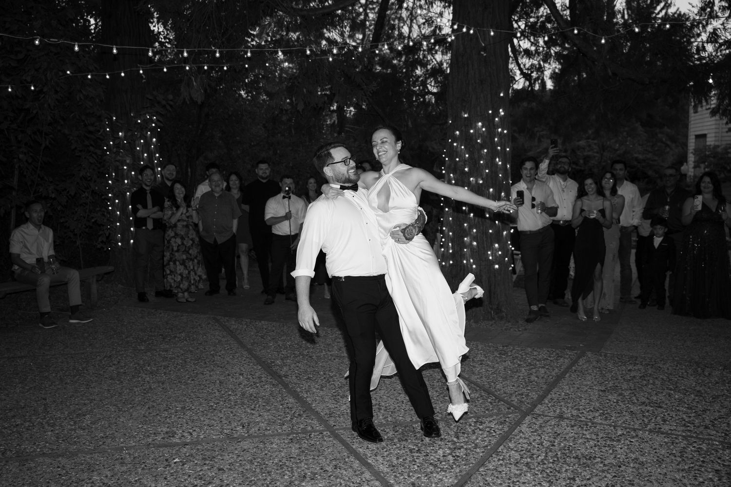 A couple dances together at an outdoor evening reception with string lights in the background.