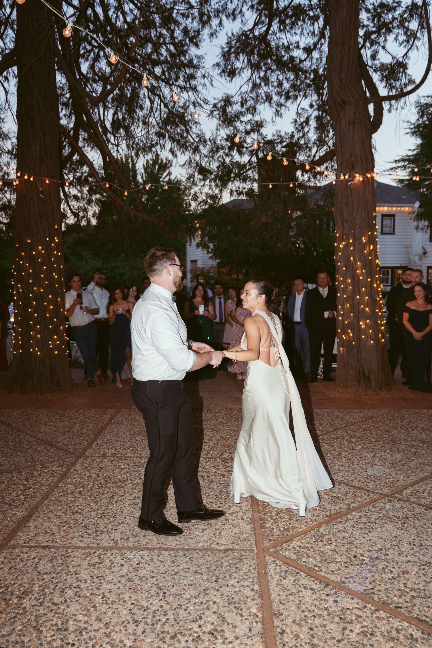 Wedding party members dance on a gravel patio decorated with twinkle lights wrapped around surrounding trees.