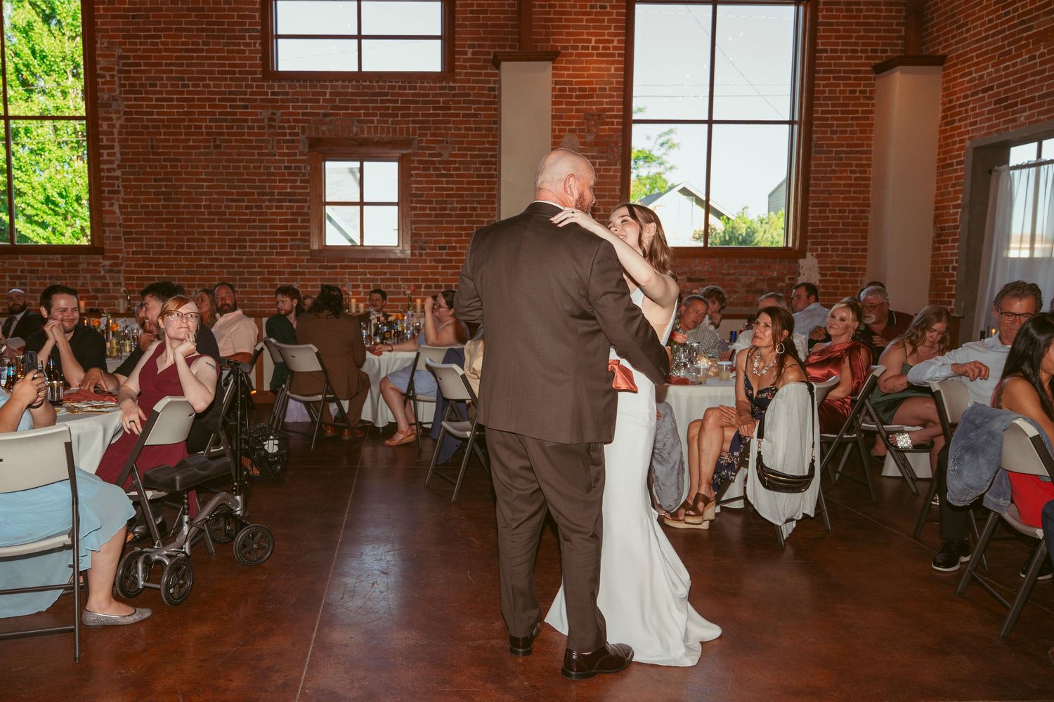 Wedding guests watch a couple dancing in an industrial-style brick venue with large windows.