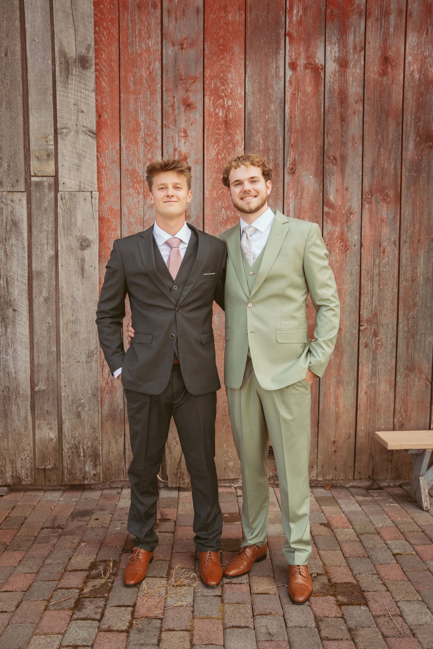 Wedding couple in contrasting suits stand together against rustic red barn backdrop.