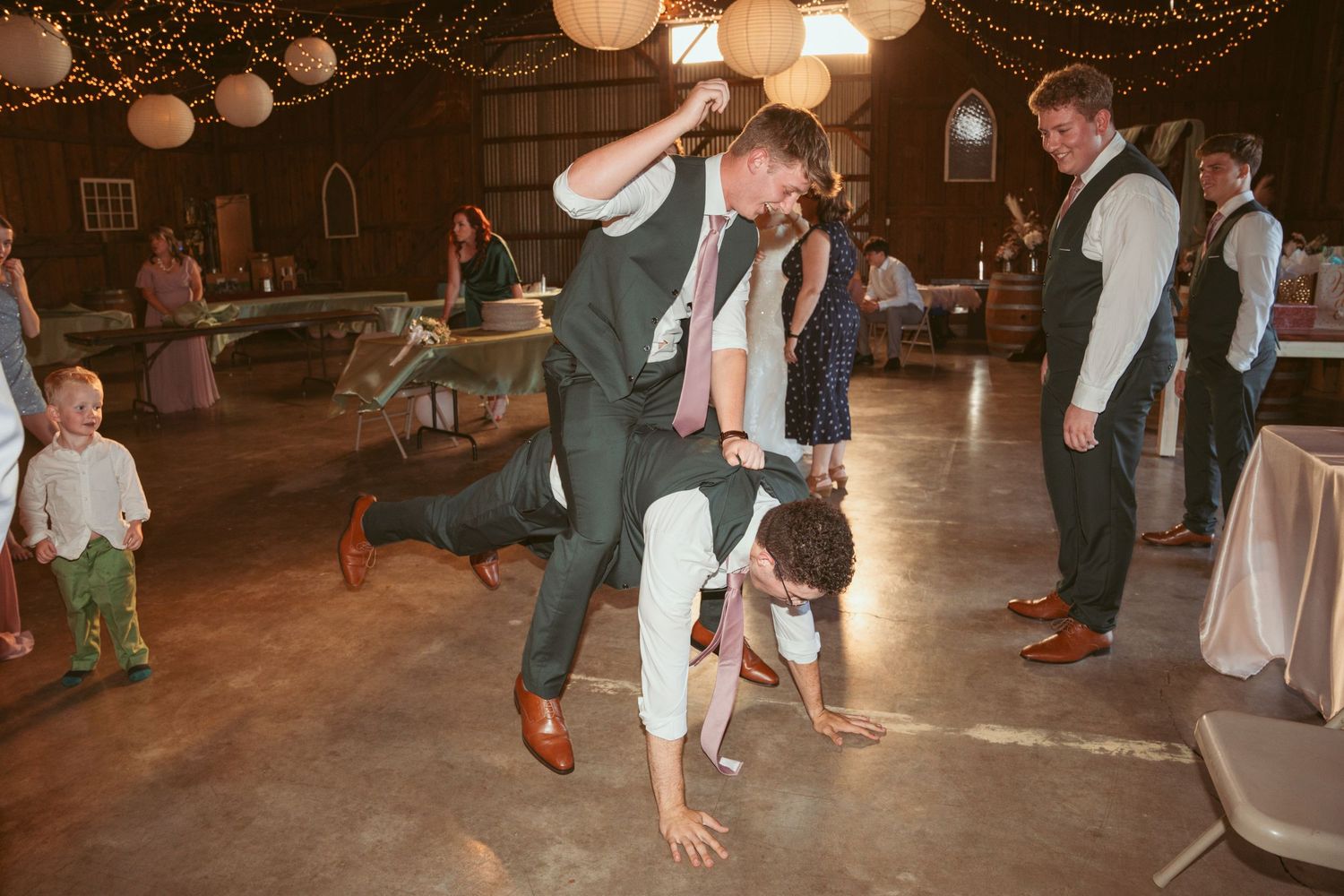 Wedding guests dance energetically under decorative string lights at an indoor reception venue.