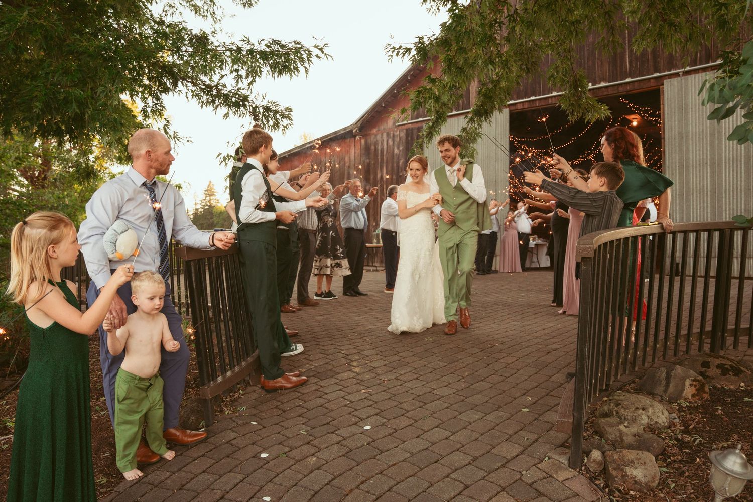 Wedding celebration outside rustic barn with guests lining wooden fence pathway.
