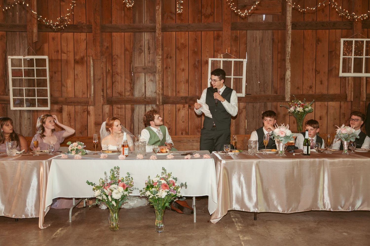 Long head table at rustic barn wedding reception decorated with pink floral arrangements and string lights.