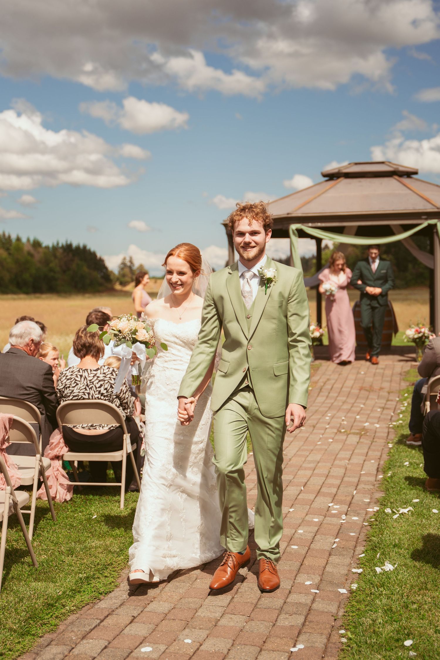 Happy couple walking down brick pathway after ceremony with gazebo and guests in background.