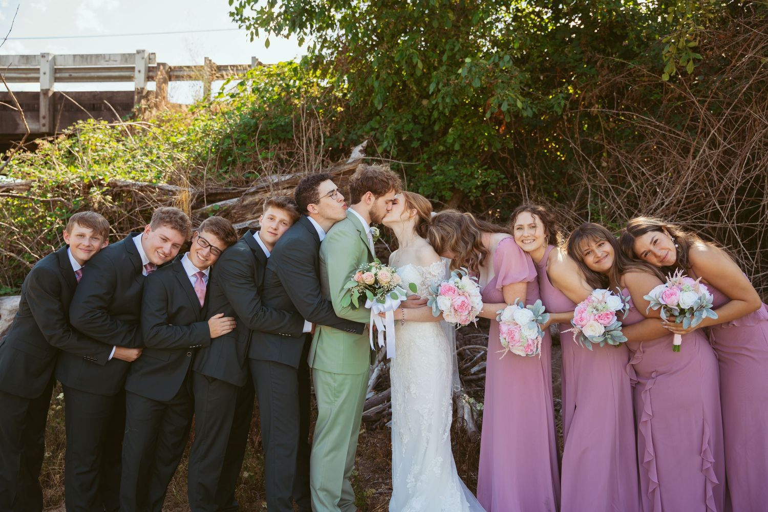 Wedding party members pose playfully in a line wearing purple dresses and dark suits.
