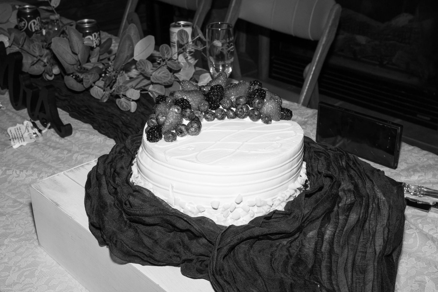 A black and white photograph of a white frosted cake with dark fabric draped around its base.