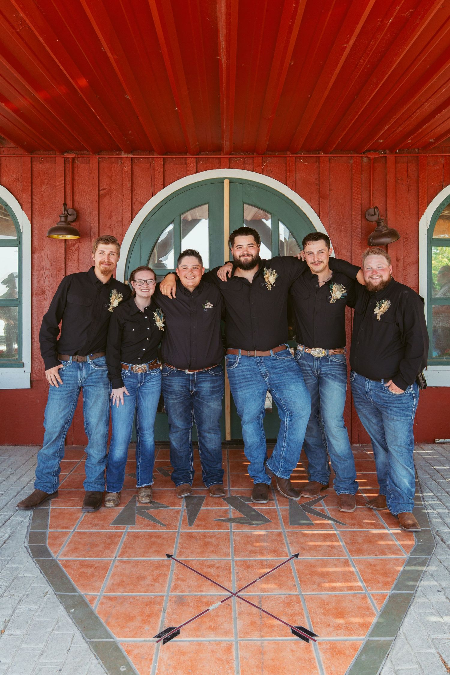 Line of groomsmen in black shirts and blue jeans pose casually against red wall with arched windows.