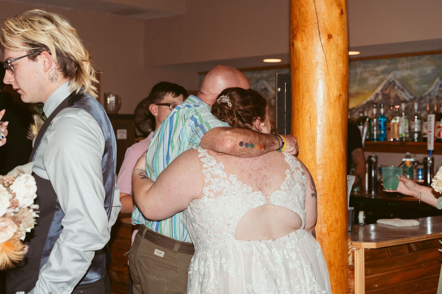 Wedding guests dancing and celebrating at a reception with wooden columns and a bar in the background.