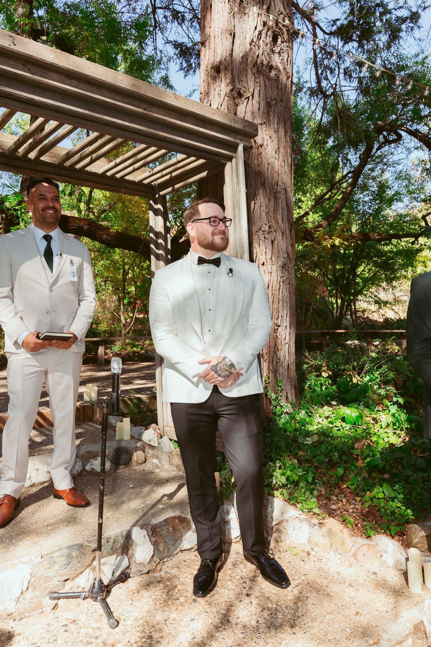 Wedding party members in white tuxedos and dark pants stand under a wooden pergola in a garden setting.
