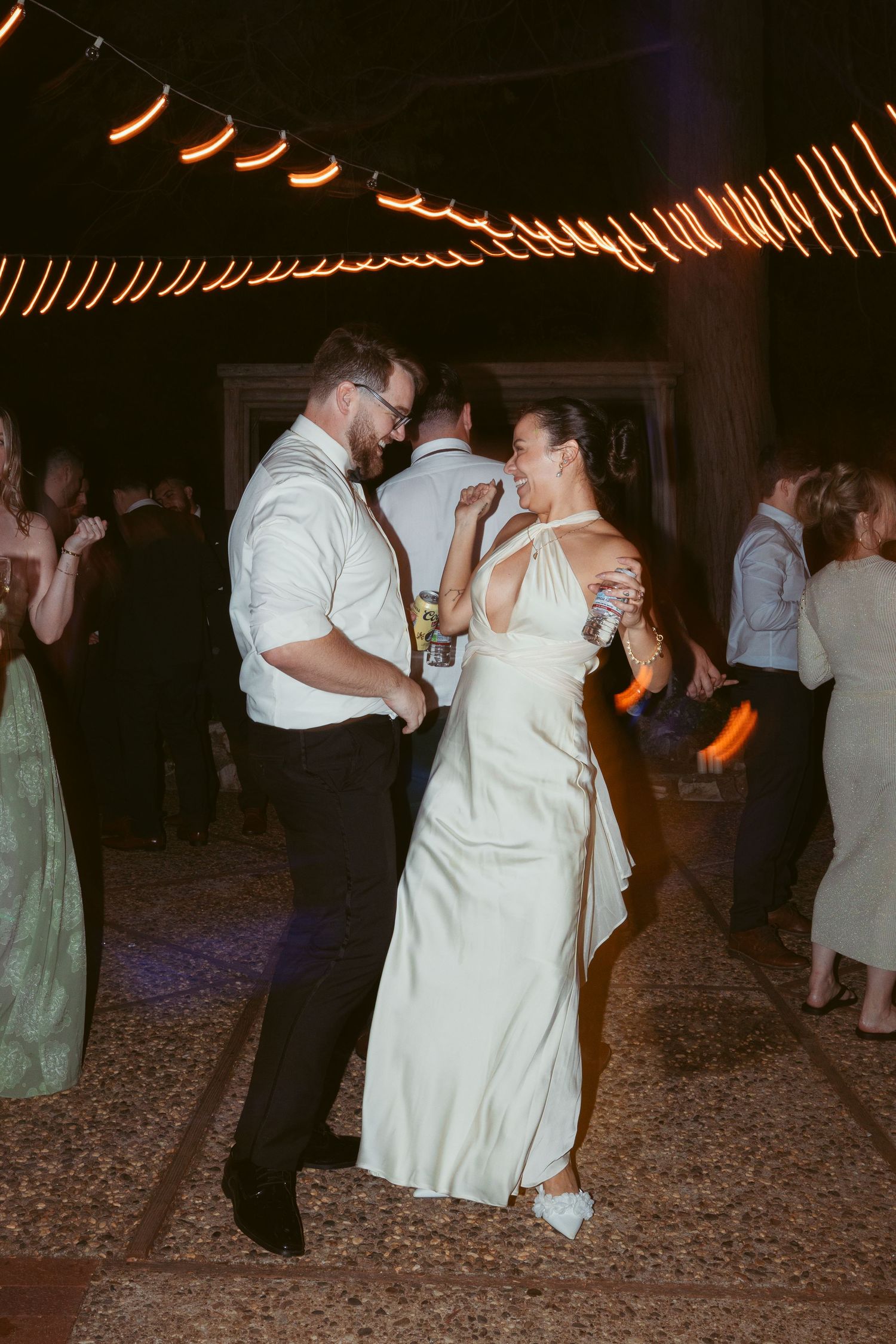 Couple dancing together at a wedding reception under warm string lights on a romantic evening.