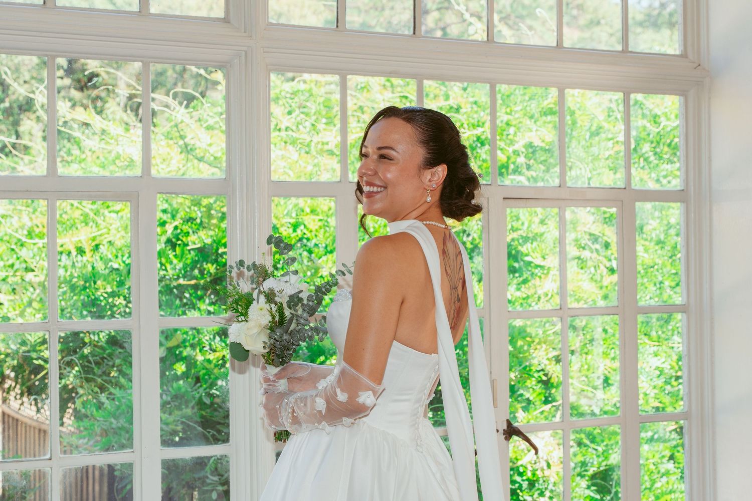 A bride in a white dress poses joyfully in a sunlit conservatory.
