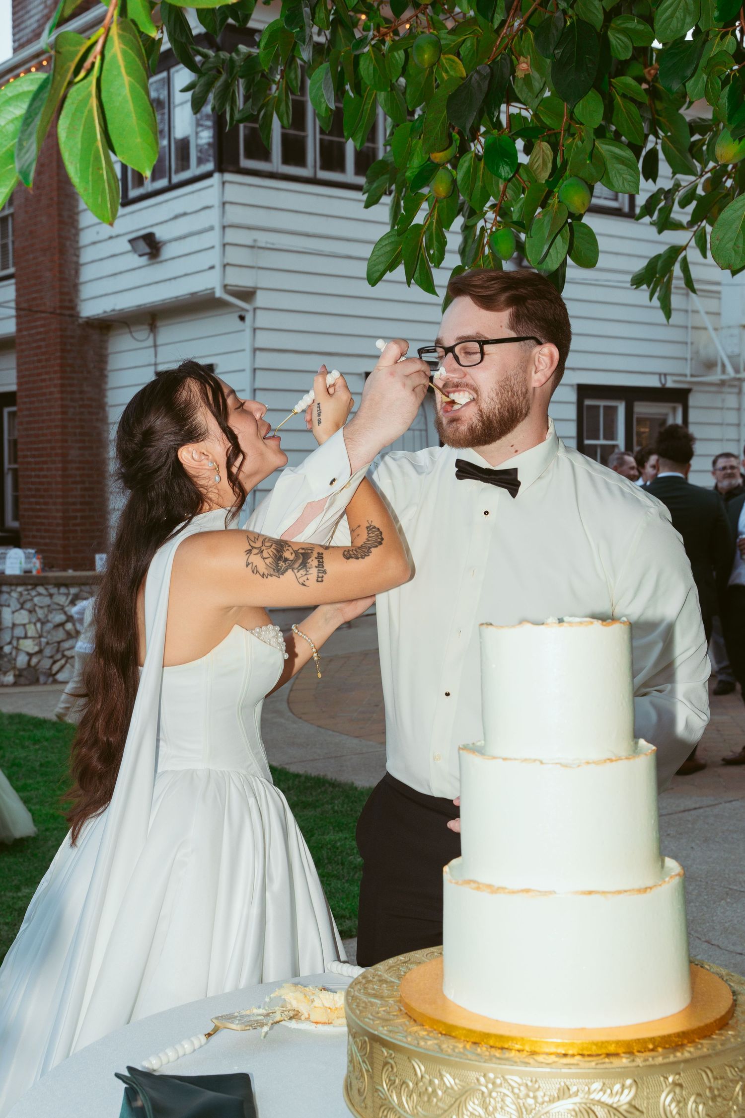 Newlywed couple playfully smashes wedding cake into each other's faces during reception celebration outdoors.