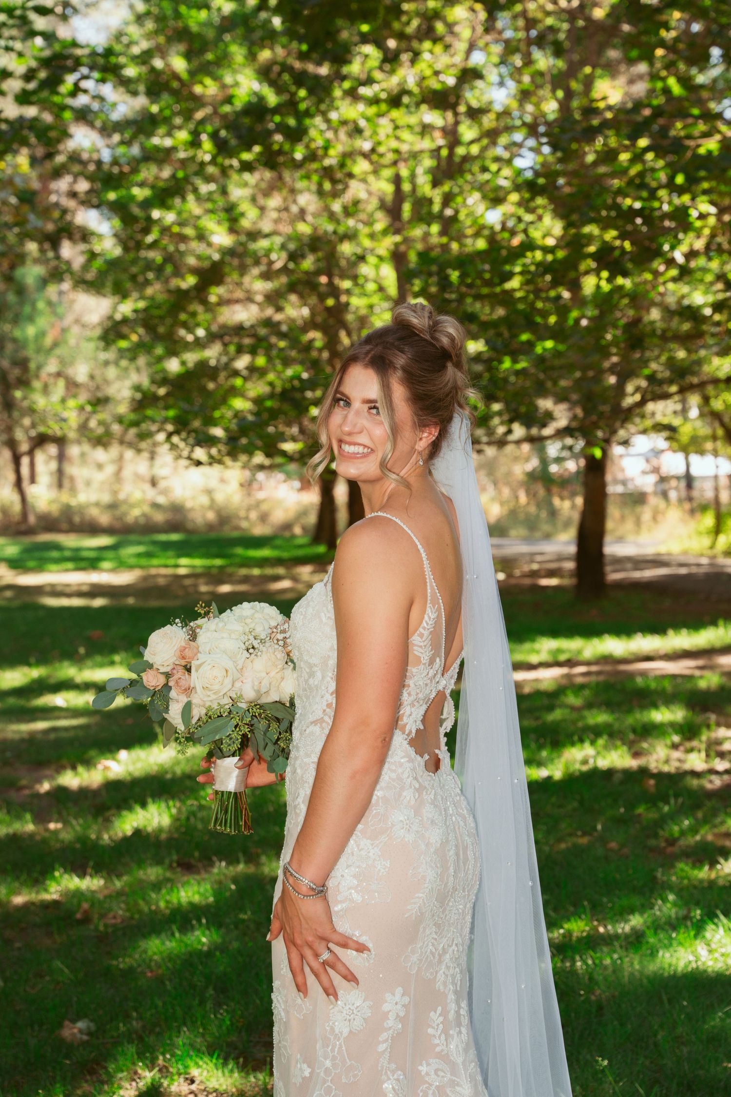 A bride in a white lace dress poses in a sunlit garden setting with a flowing veil and bouquet.