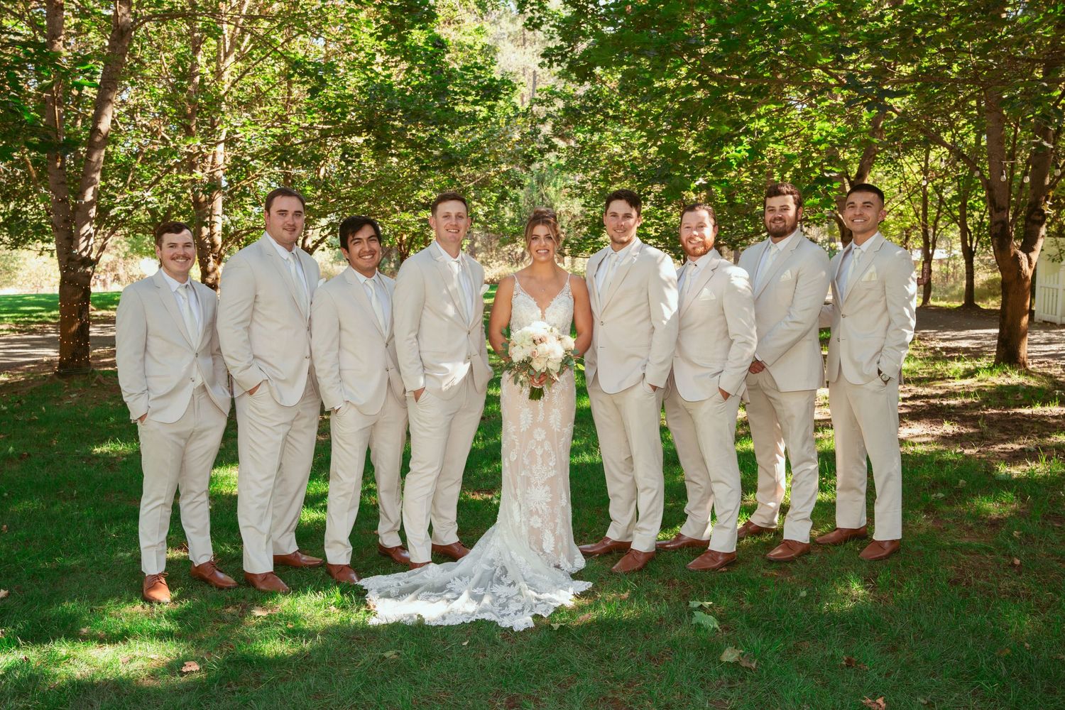 Wedding party poses in coordinated white suits with person in white gown centered in outdoor garden setting.
