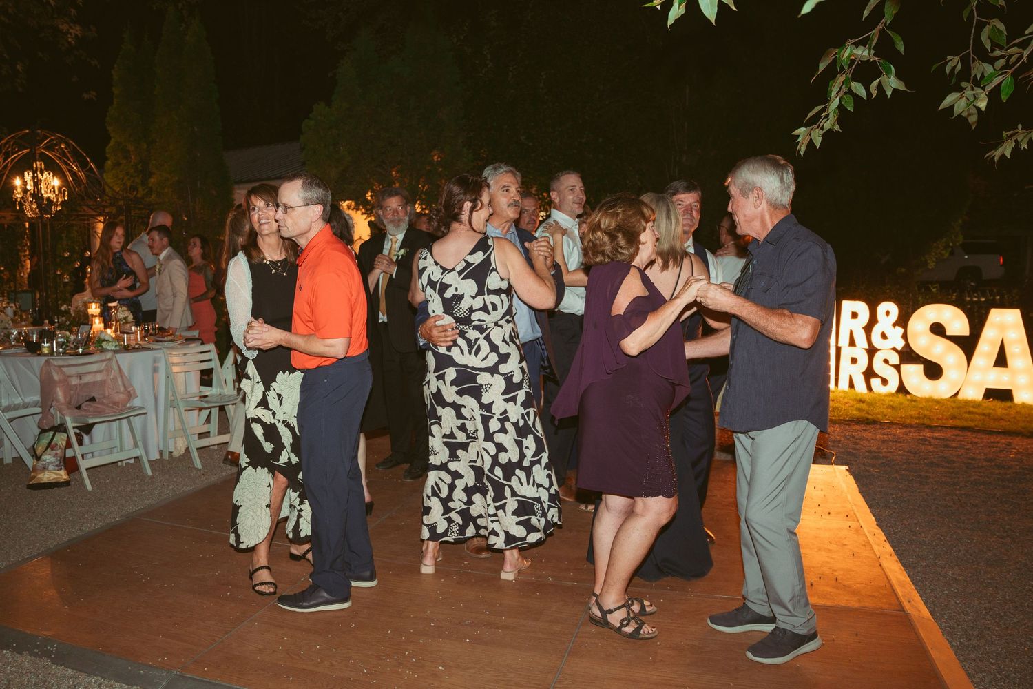 Wedding guests dancing in pairs on an outdoor dance floor with illuminated signage visible.