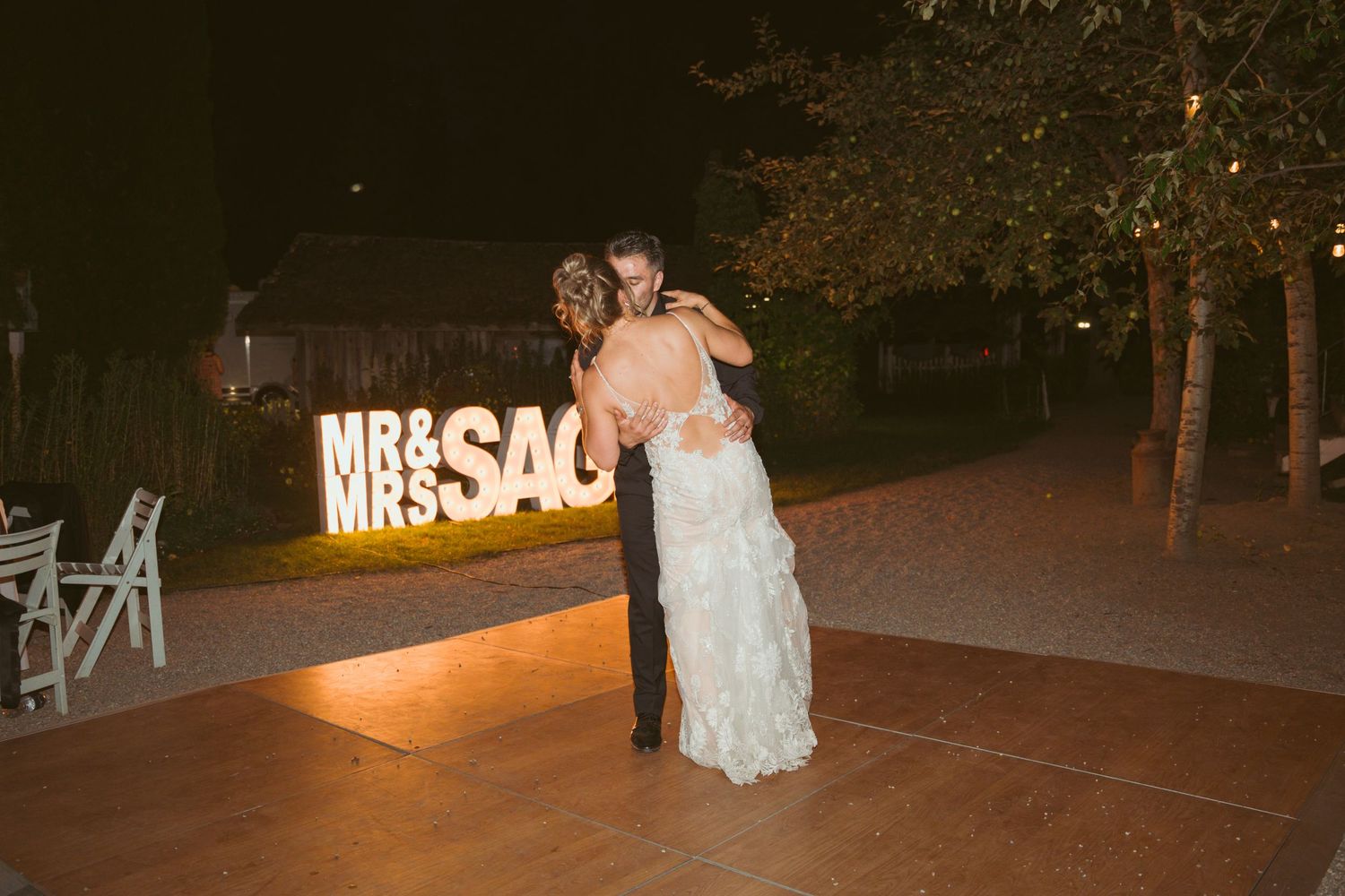 Evening wedding reception with wooden dance floor and lit signage creating a romantic atmosphere.