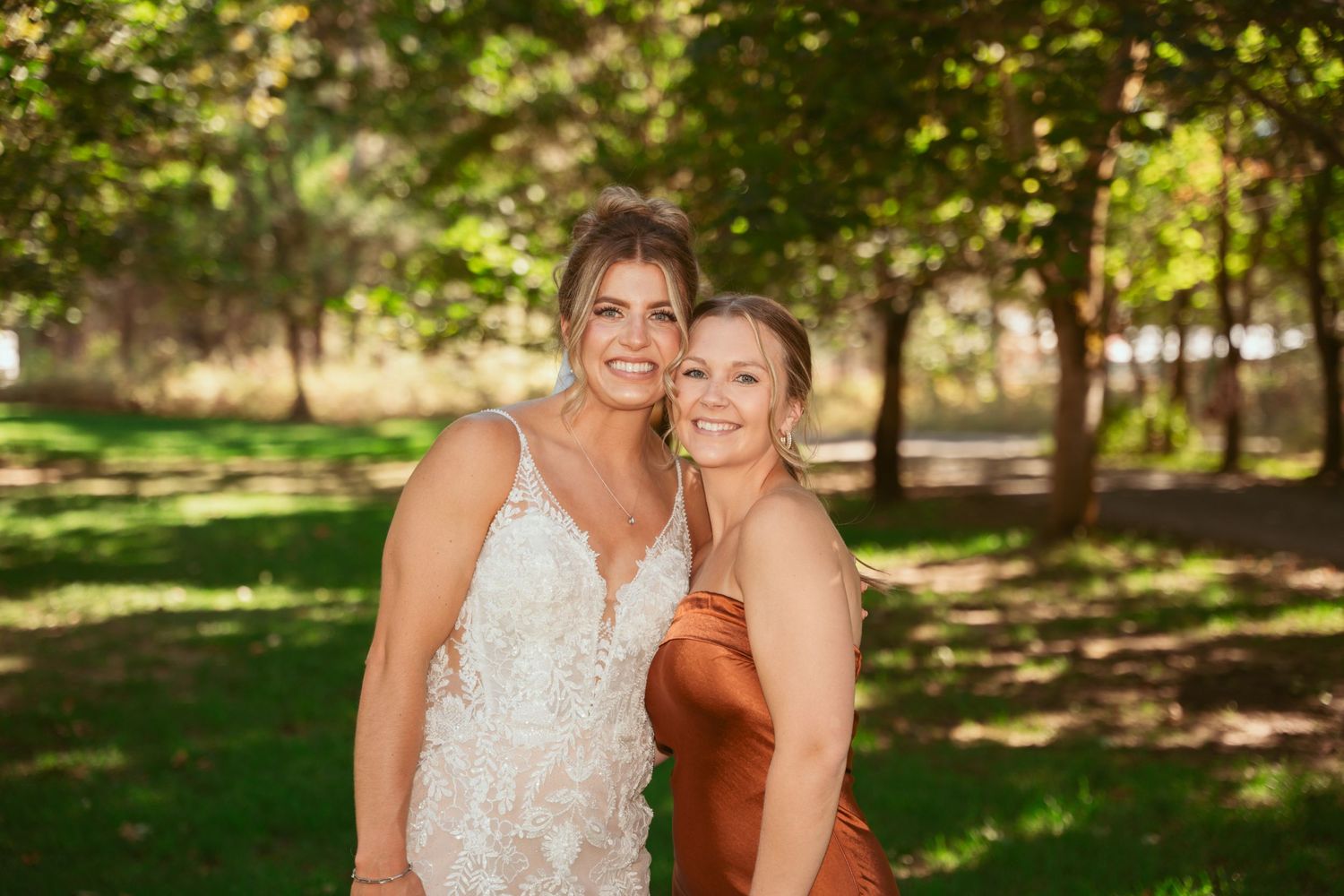 Two people in formal wear share a close moment outdoors under green trees.