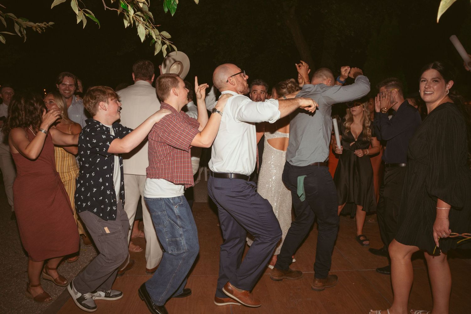 Large group of wedding guests dancing in a line during an evening outdoor reception.
