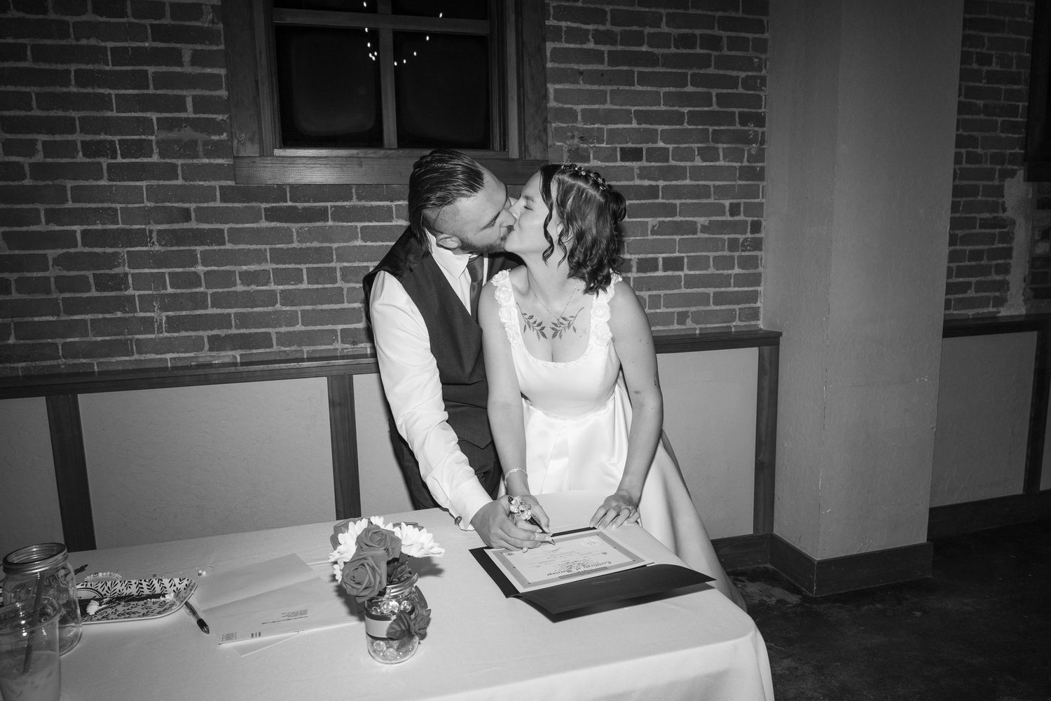 Couple signing documents at table during wedding reception in brick-walled venue.