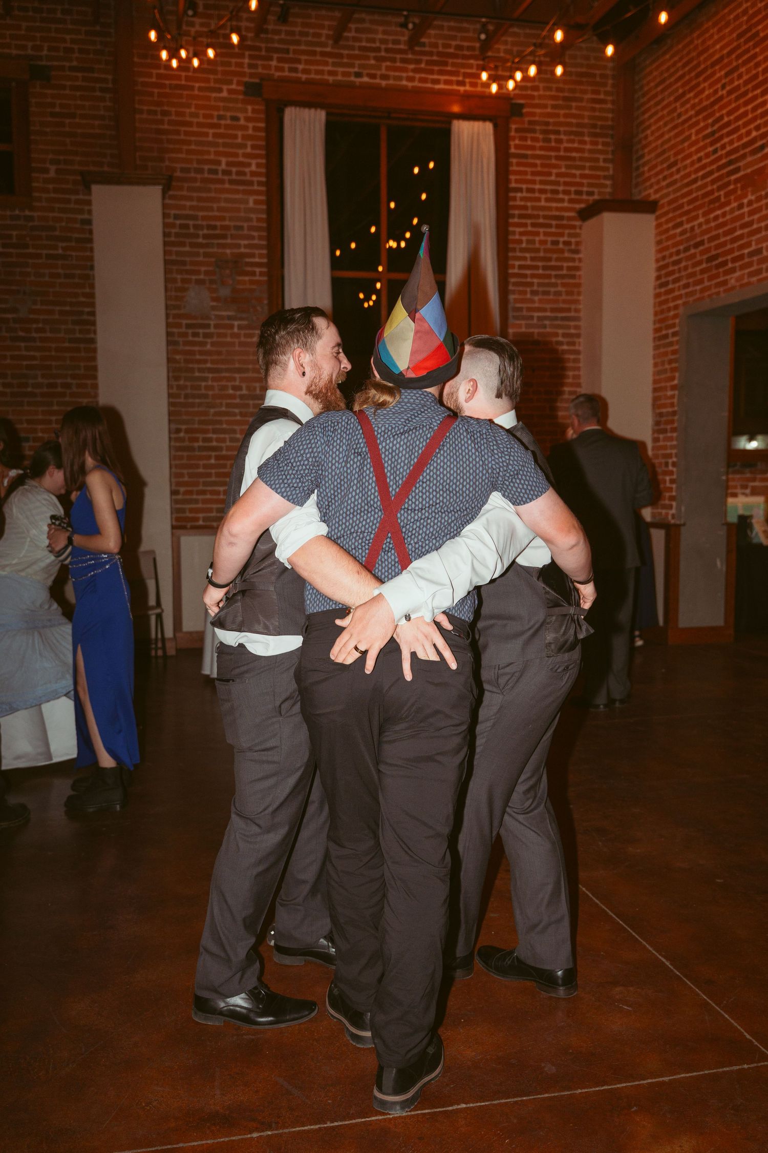 Wedding party members in dark suits dancing in formation under string lights.