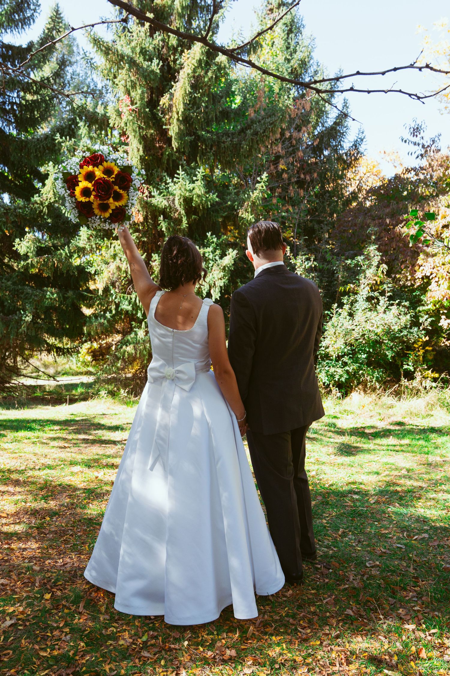 A couple stands in a forest clearing with the bride holding up a sunflower bouquet.
