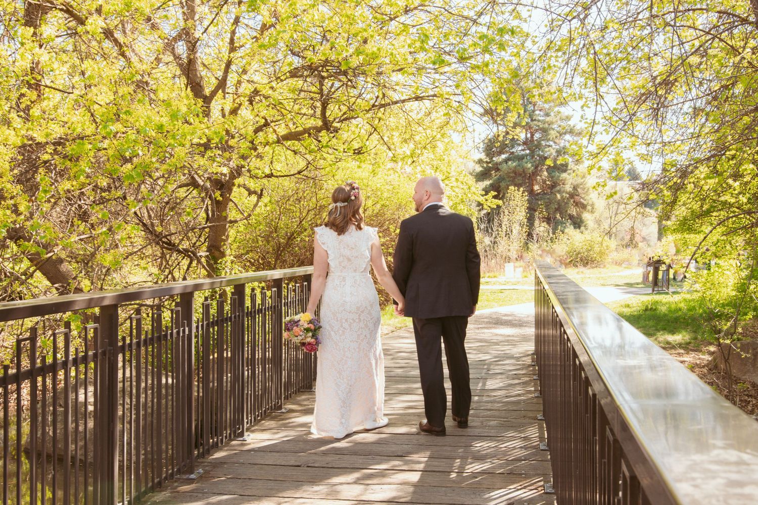 Wedding couple walks along scenic iron-railed pathway with trees and water features in background.