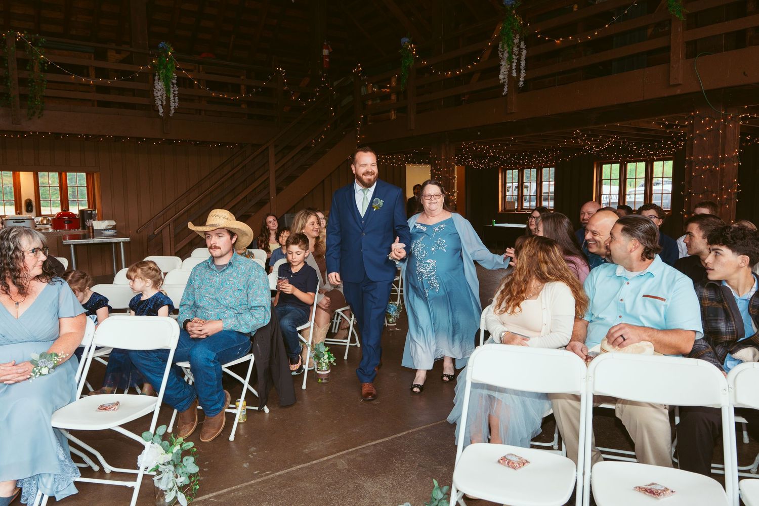 Wedding celebration in a spacious barn venue with white folding chairs and string light decor.