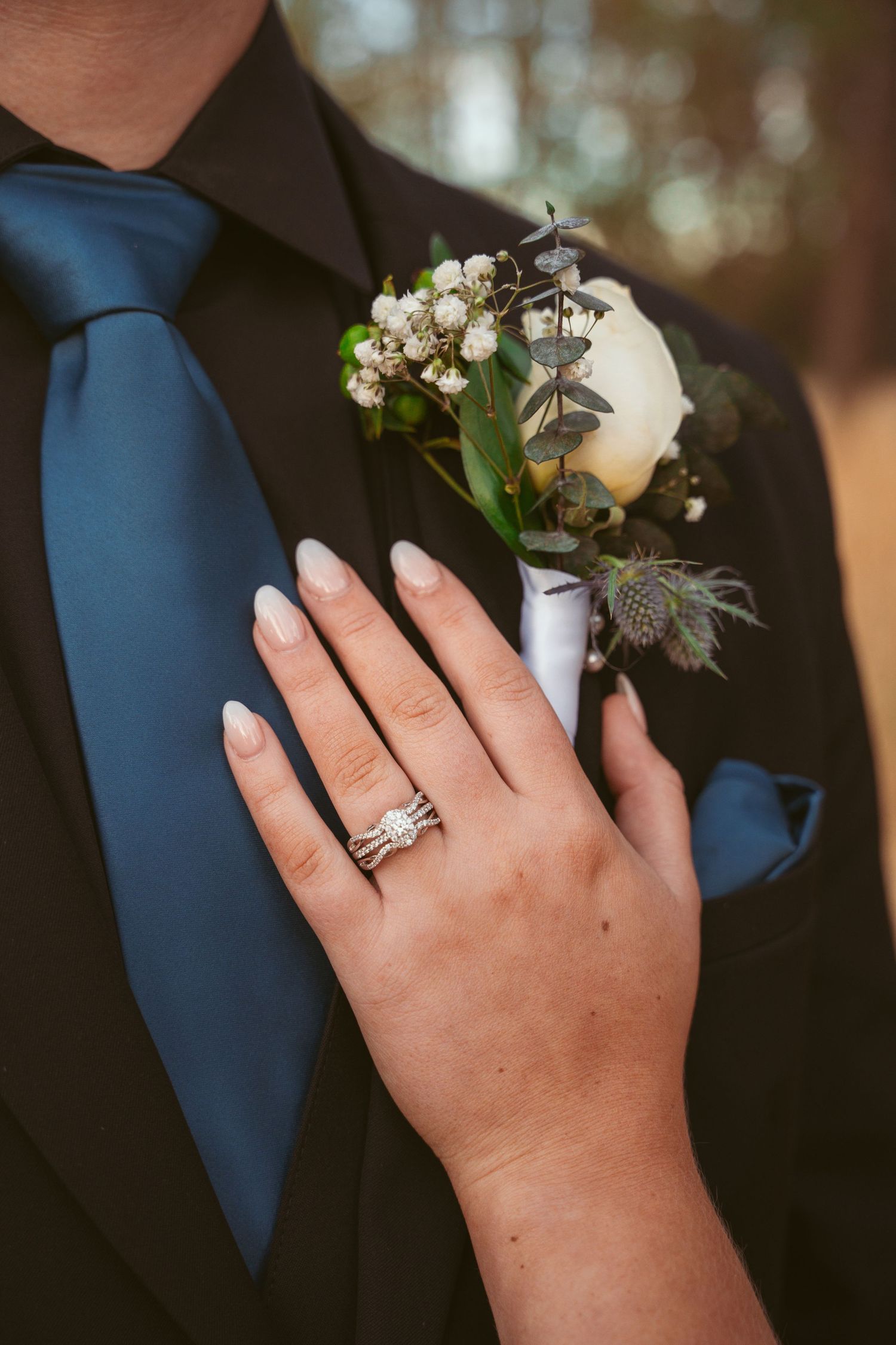 Close-up of a hand wearing wedding rings touching a boutonniere with baby's breath and greenery against a navy blue tie.