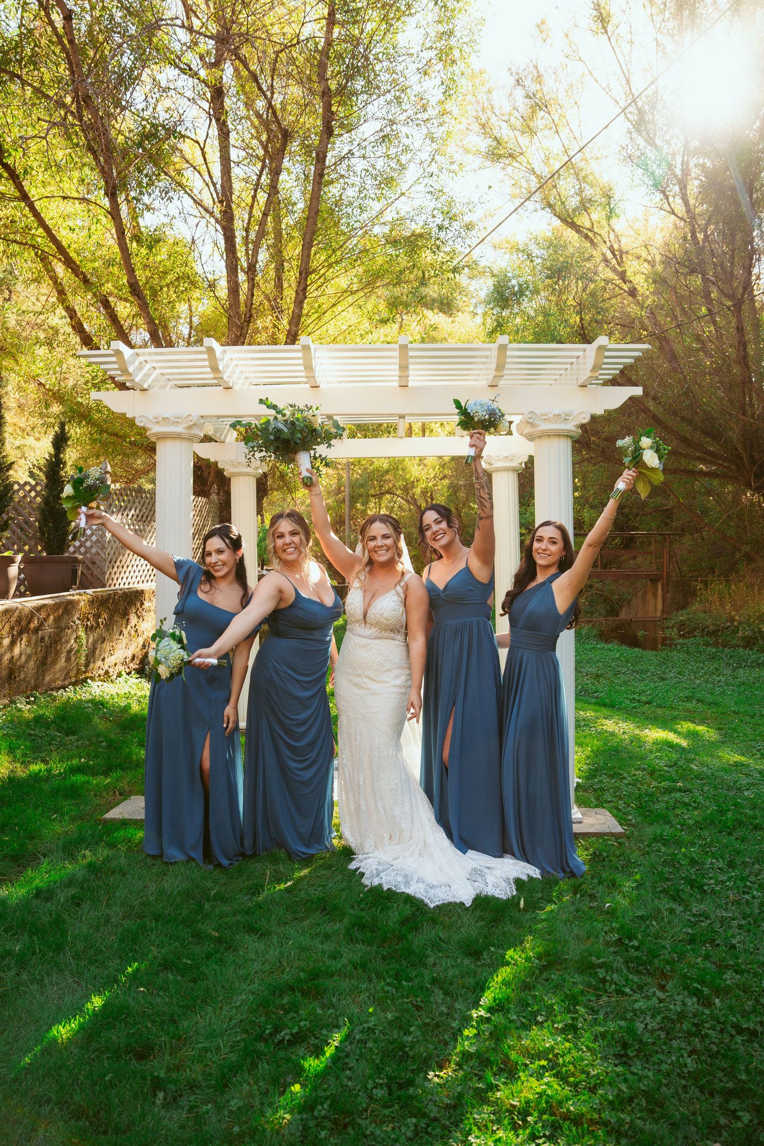 Wedding party in navy blue dresses poses playfully on green lawn with white pergola backdrop.