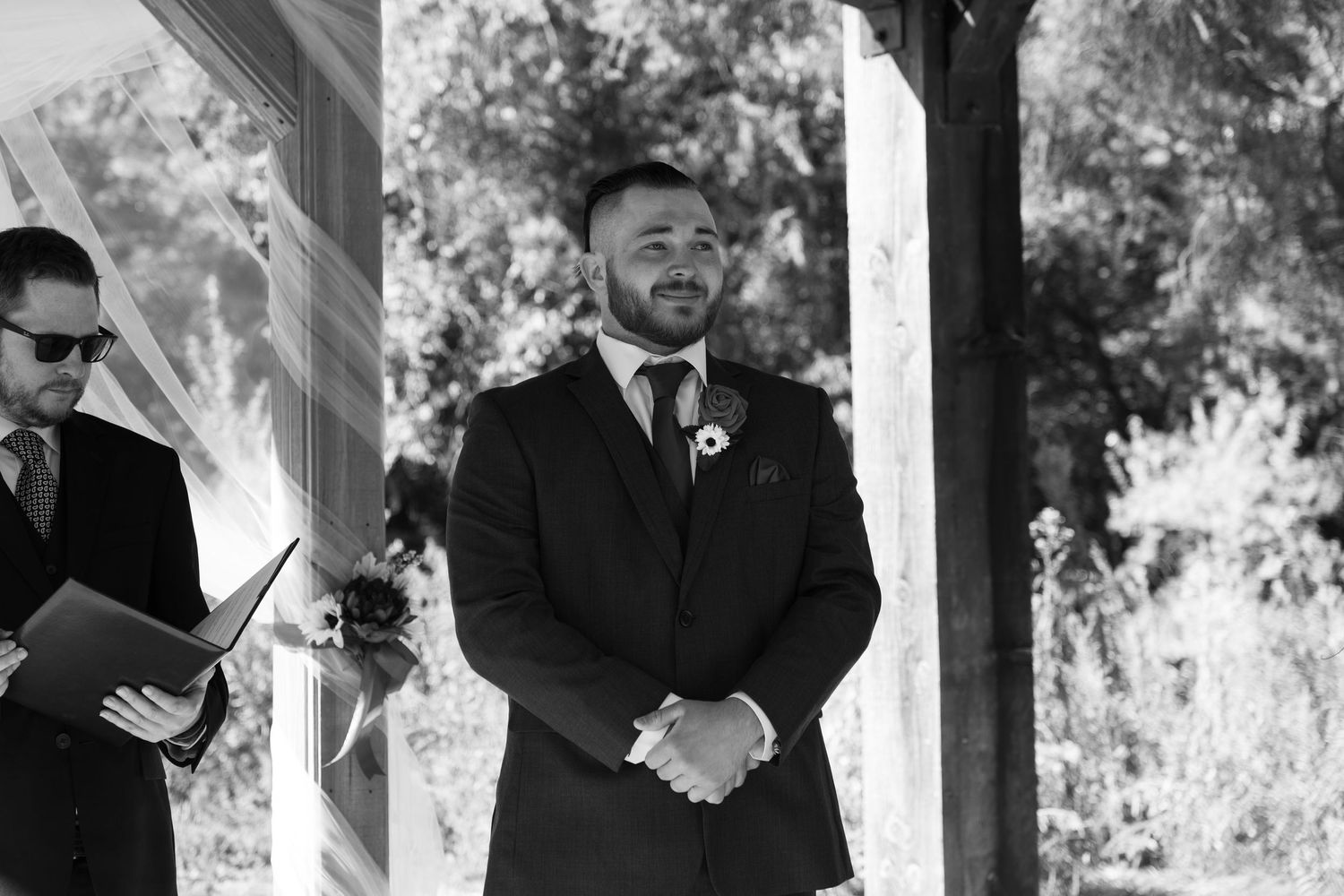 Black and white photo of groomsmen standing under rustic wooden pergola during ceremony.