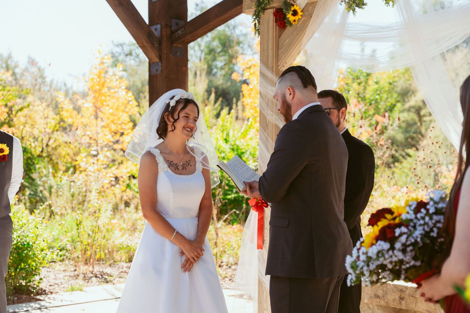 Outdoor wedding ceremony under a rustic wooden pergola decorated with sunflowers and fall foliage.