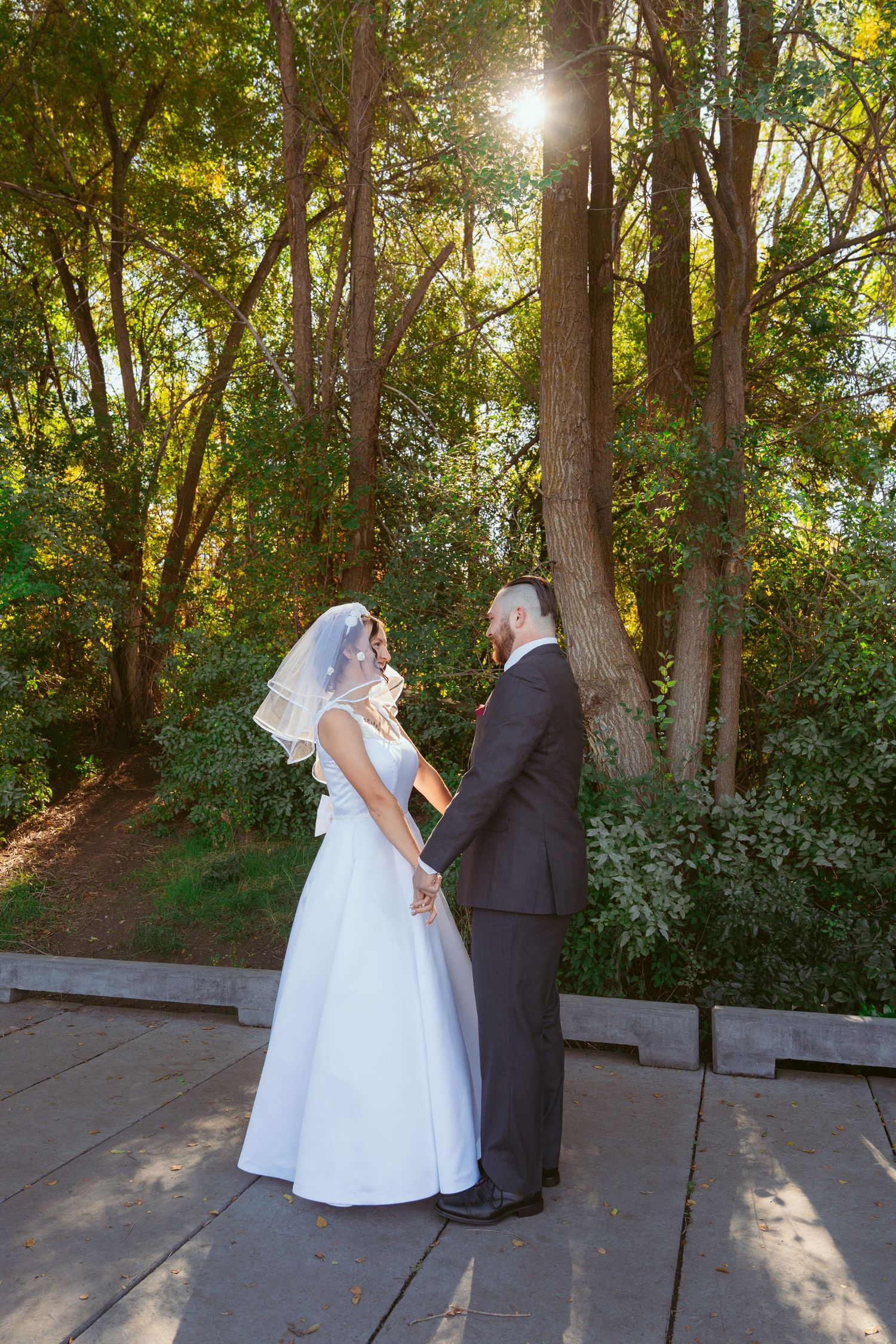 Wedding couple shares romantic moments on wooden deck with sunlight streaming through forest trees.