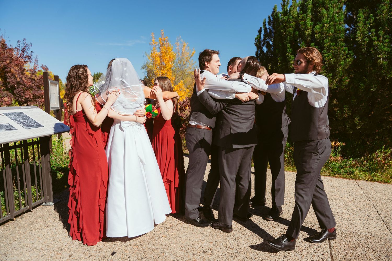 Wedding party lined up outdoors against autumn trees and blue sky.