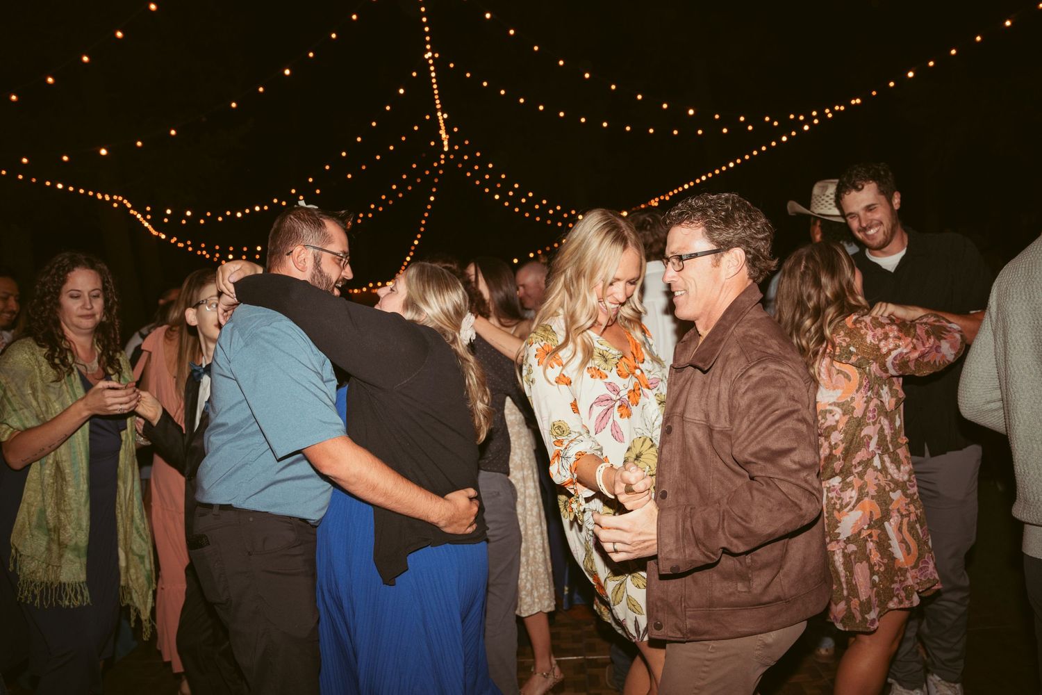 Wedding party dancing and celebrating under string lights at an outdoor evening reception.