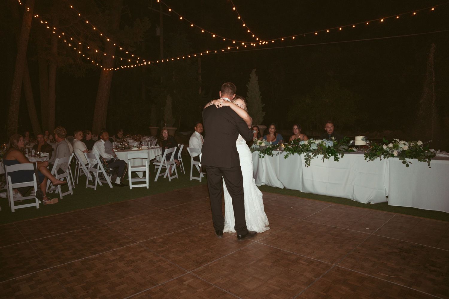 Wedding guests dance on a rustic wooden dance floor under glowing string lights at an outdoor evening reception.