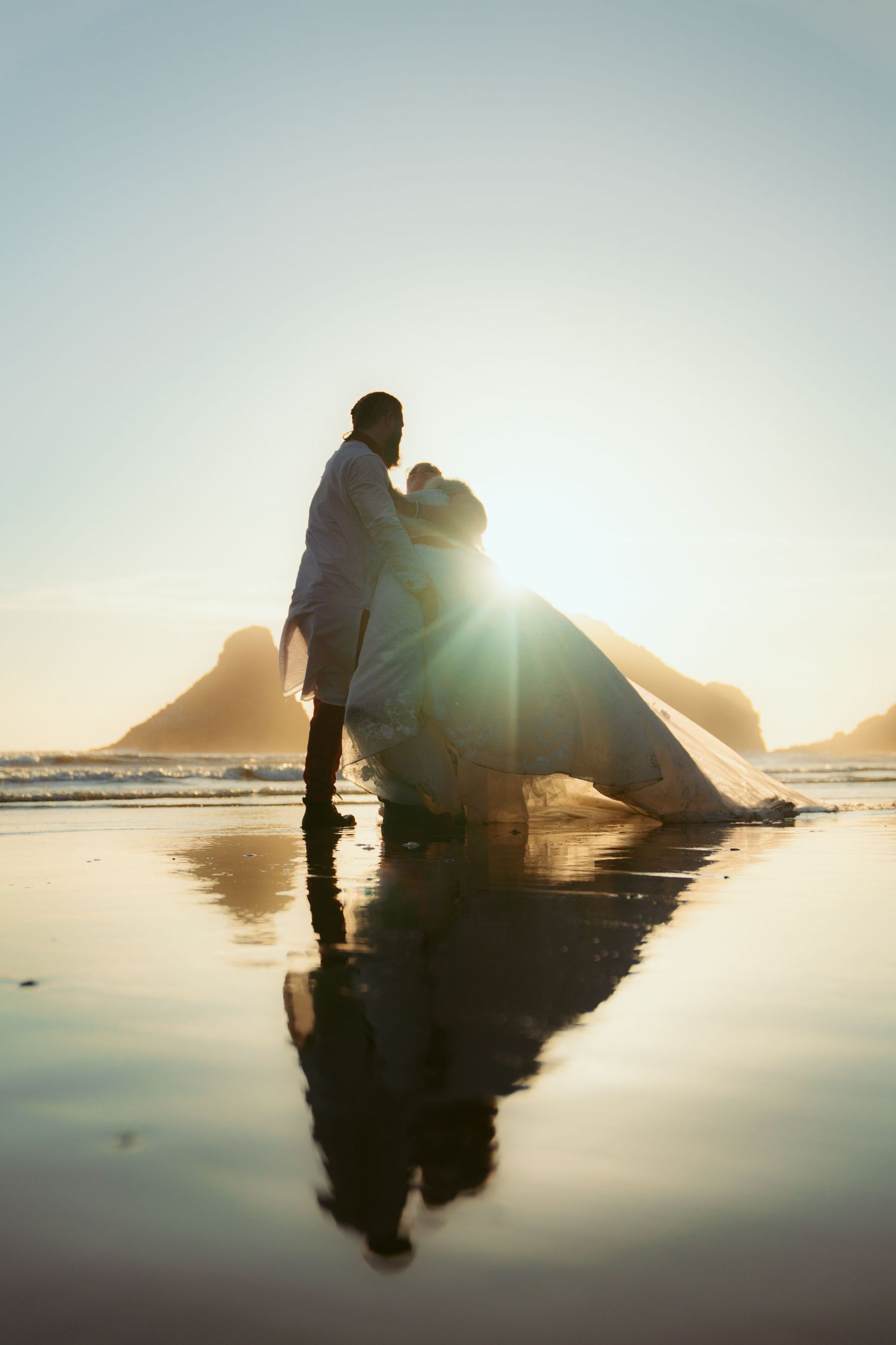 Romantic silhouettes of a couple on a beach at sunset with reflections in wet sand and mountains in background.