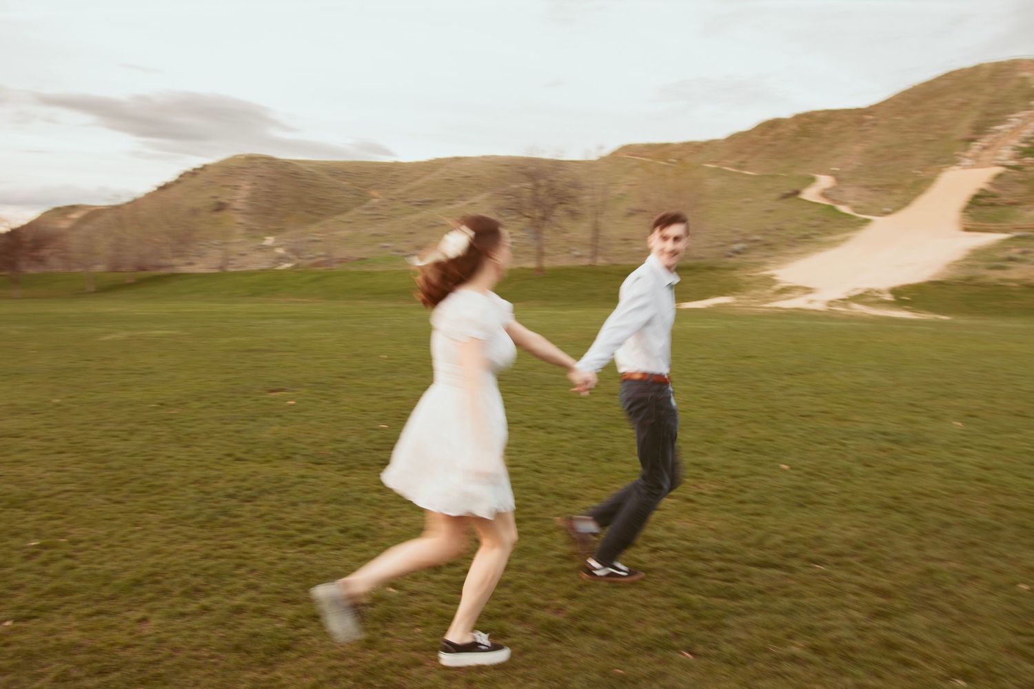 Two people dance and spin joyfully across an open grassy field with mountains in the background.