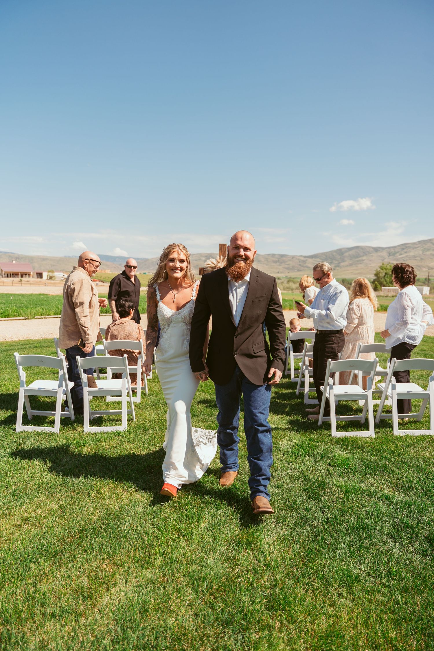 Wedding guests gather on white chairs on a grassy lawn with mountains in the background.