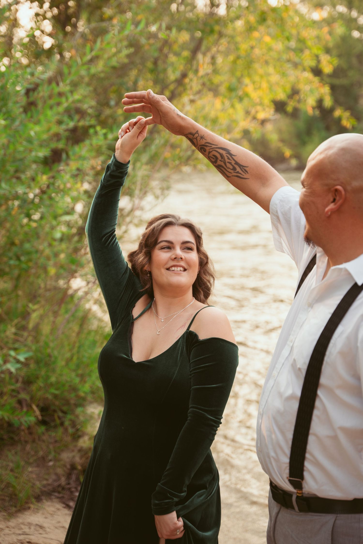 A couple dances joyfully on a sunlit forest path during an outdoor photoshoot.