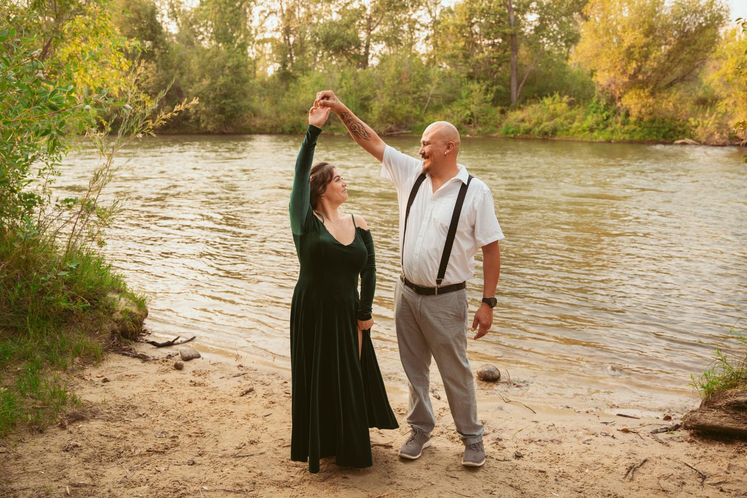 Romantic couple posing playfully by a riverbank during golden hour with autumn foliage in the background.