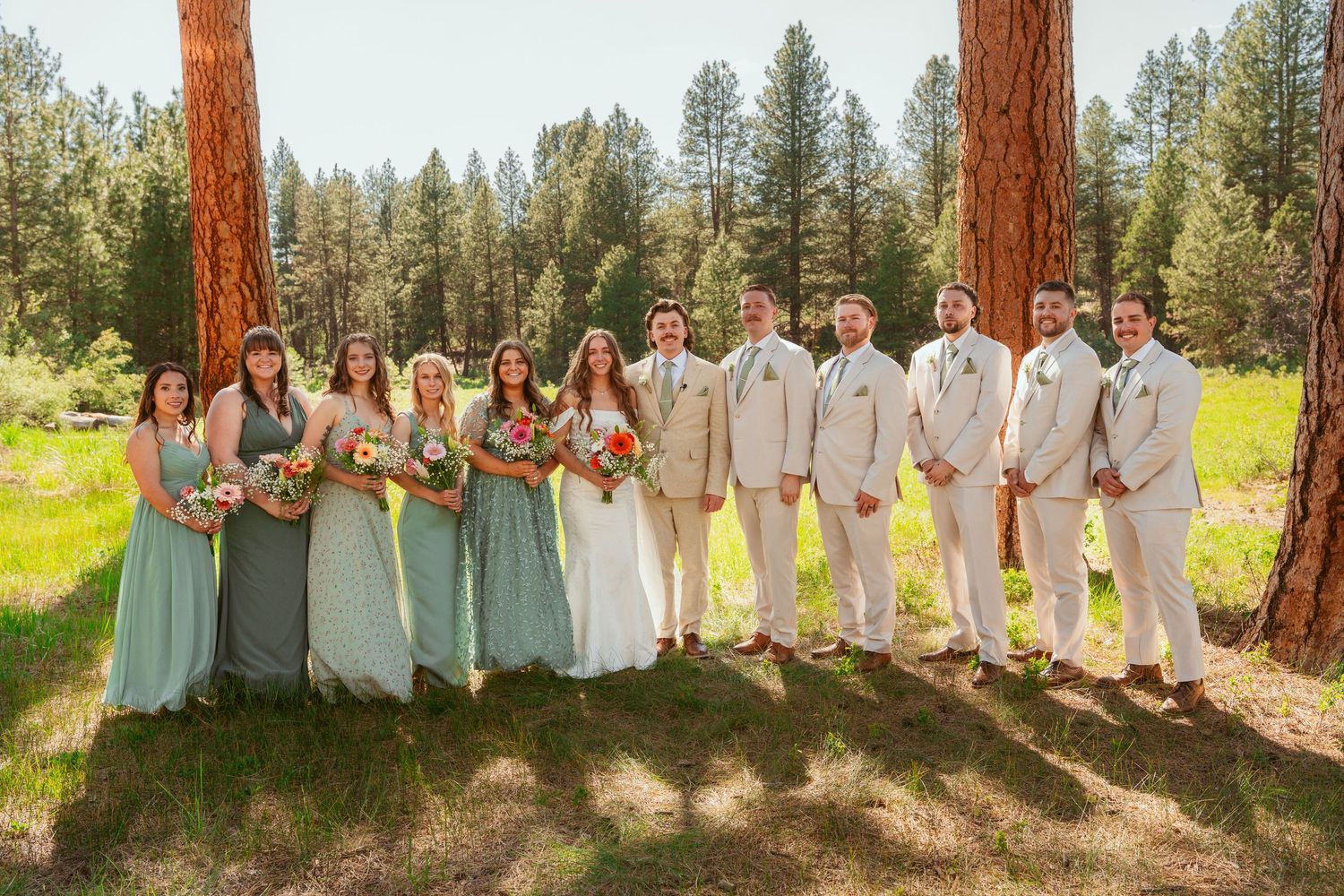 Large wedding party in coordinated sage and white attire poses in forest clearing.