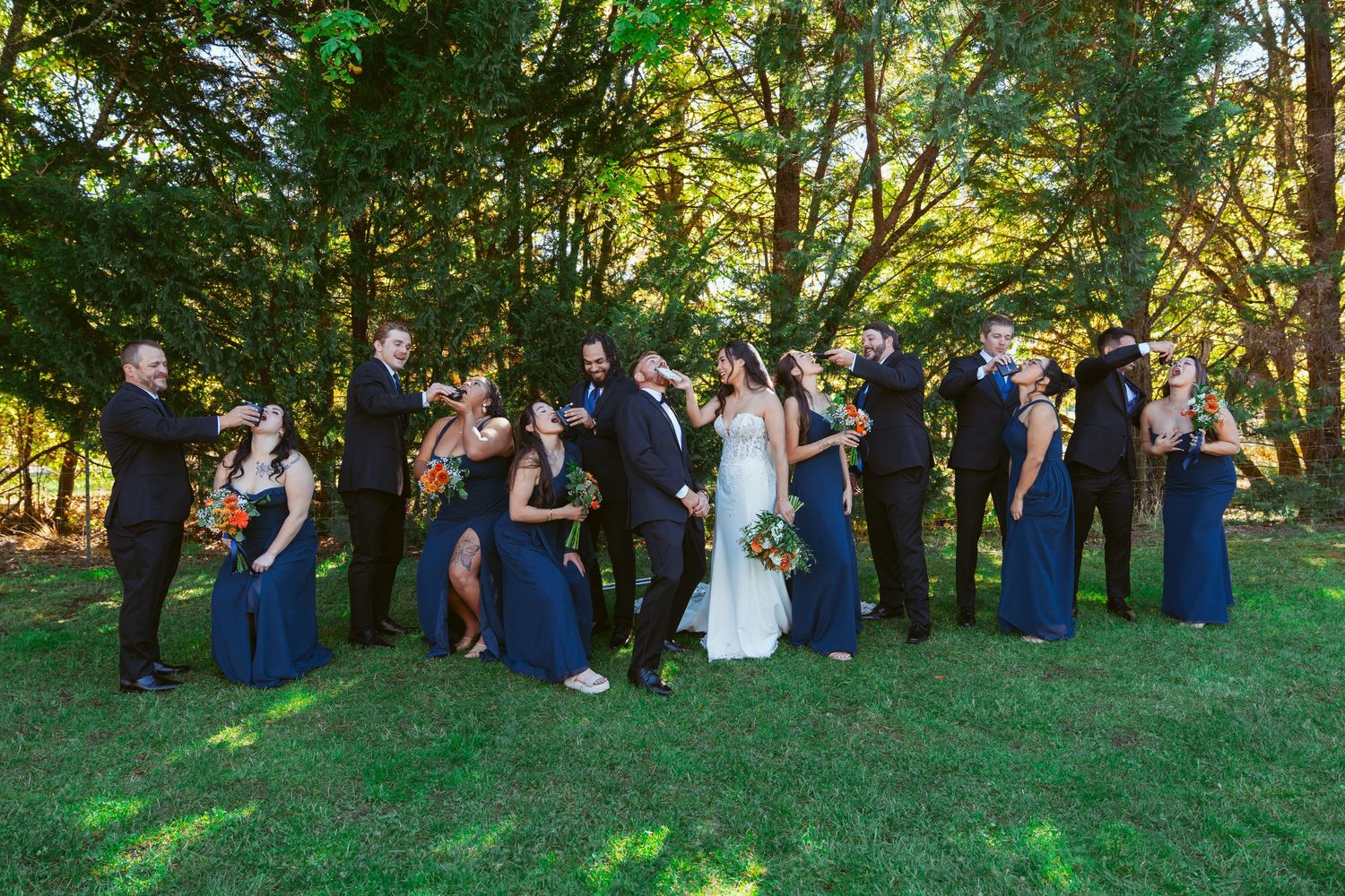 Wedding party members line up on grassy hill for panoramic group photo in navy and white attire.