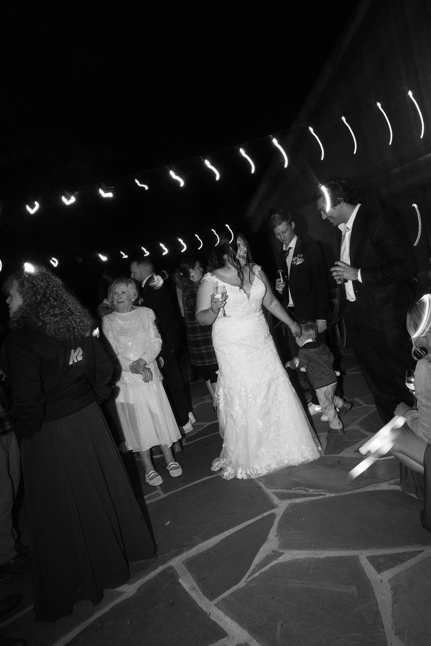 Wedding guests dance together on a dark dance floor with raised hands during an evening reception celebration.