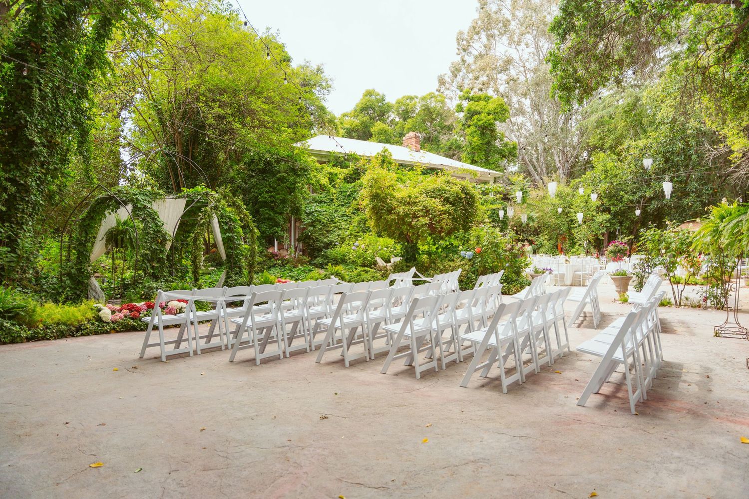 White ceremony chairs arranged in rows on a garden patio surrounded by lush greenery.