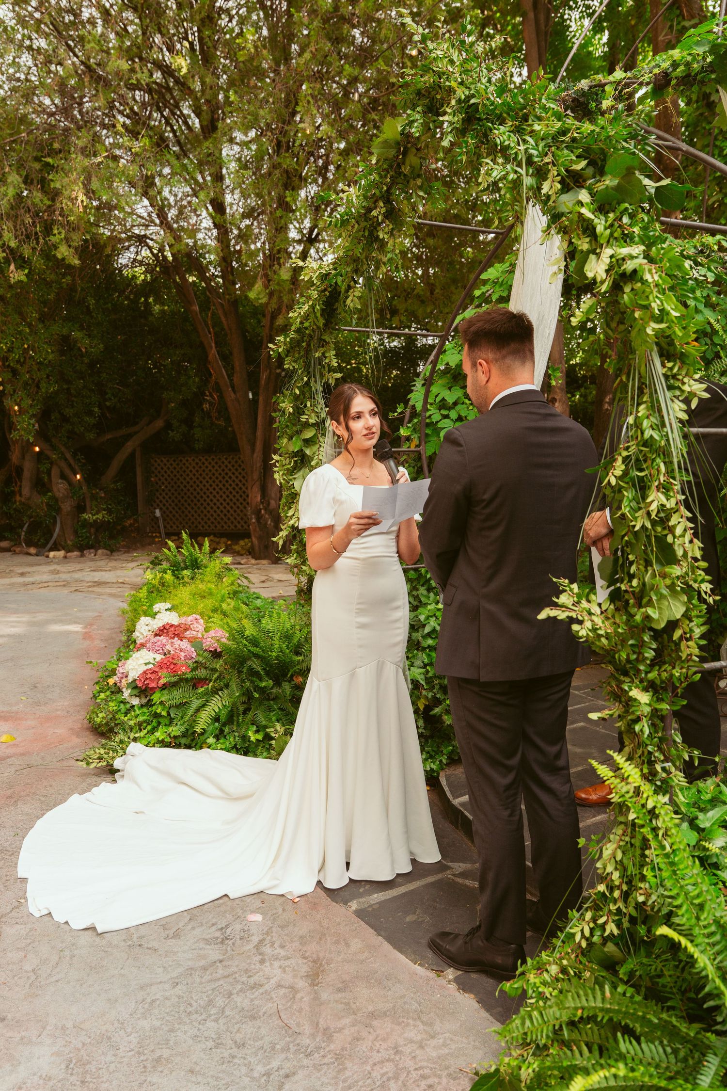 Wedding couple exchanging vows under a canopy of trees with white decor and floral arrangements.